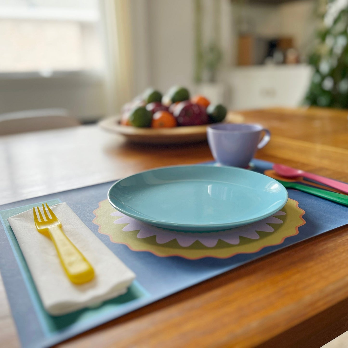 Teal plate on a colorful placemat with cutlery on a wooden table.