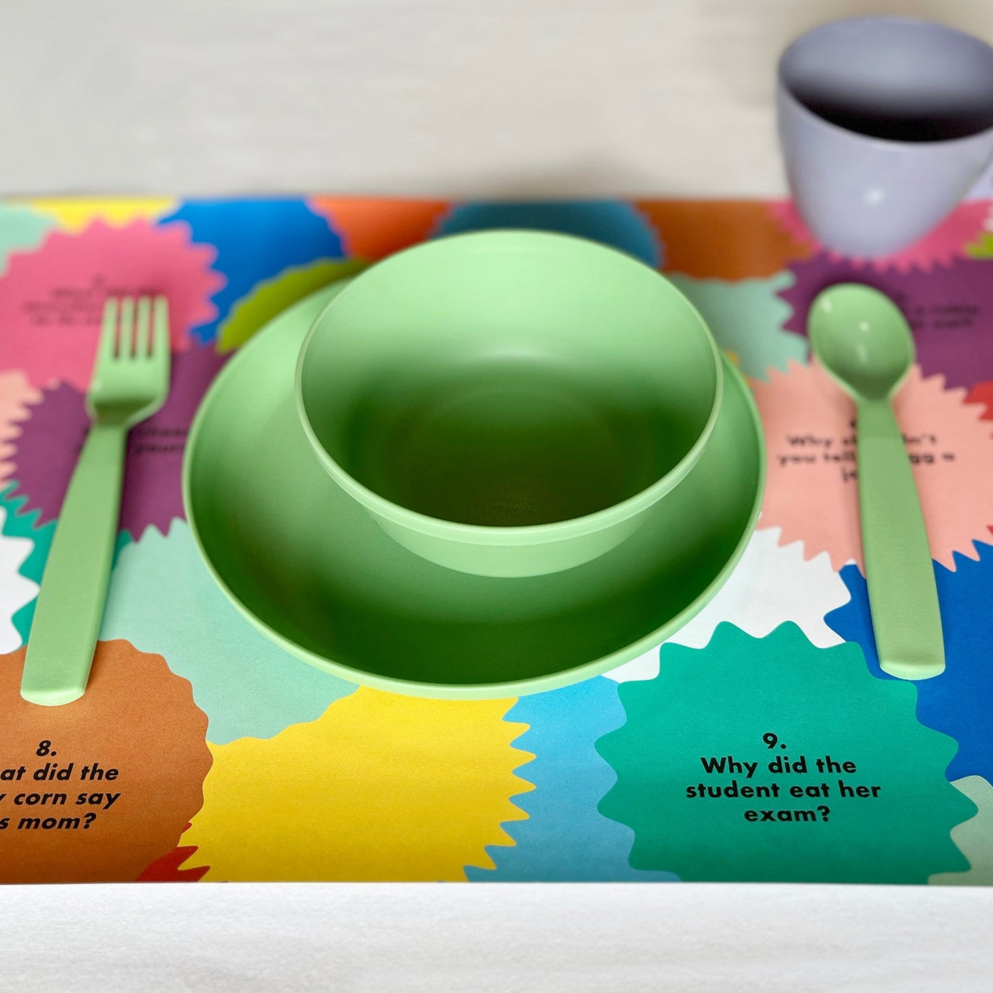 Colorful paper placemat with green bowl, fork, and spoon on a white background