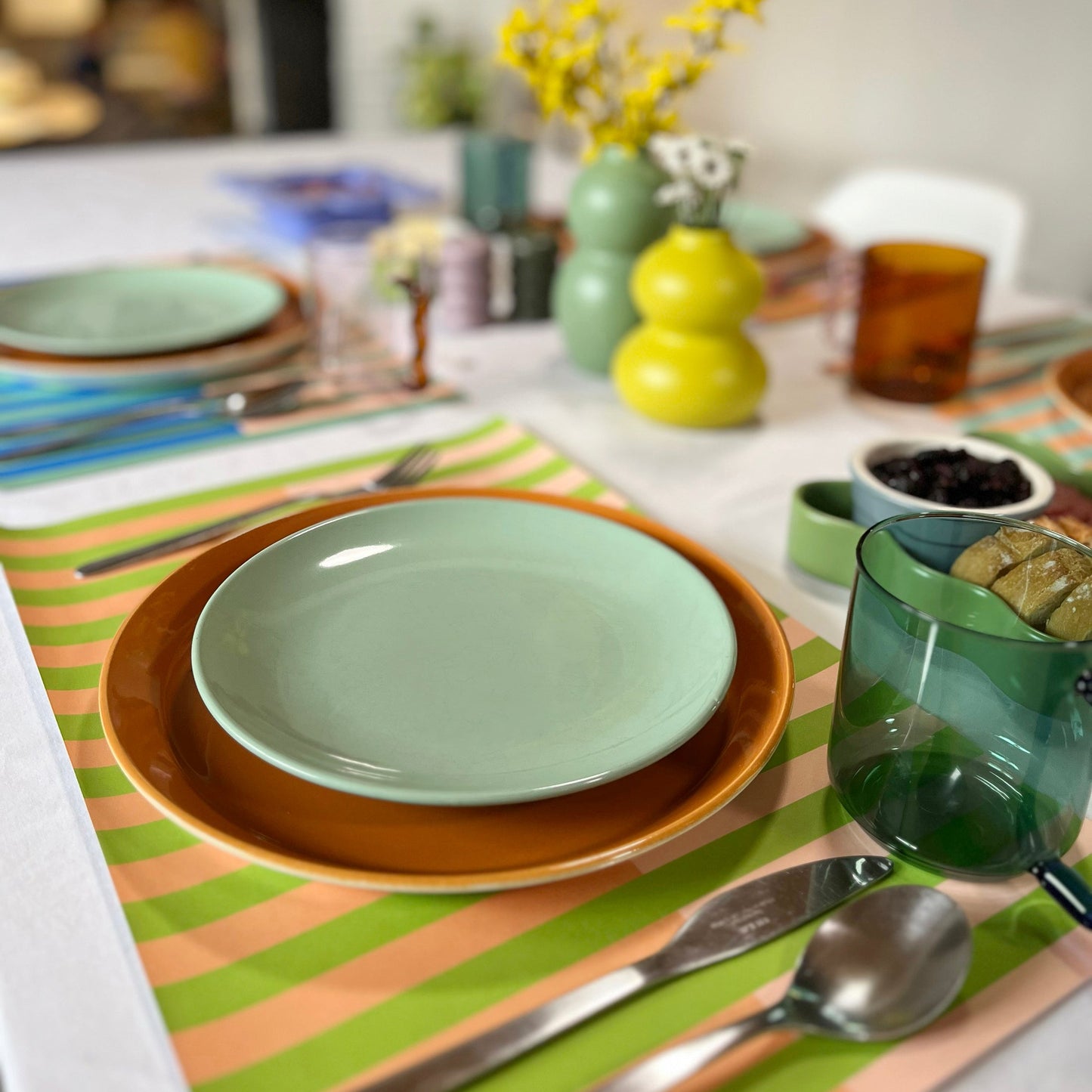 Dining table setting with green plates, glasses, and colorful striped paper placemats.