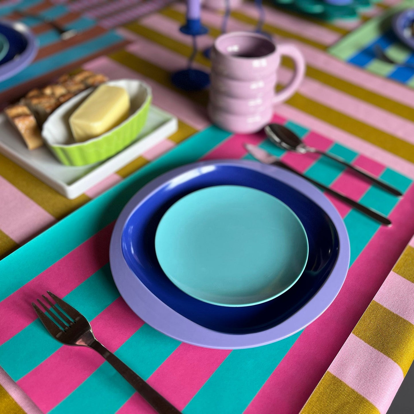 Colorful table setting with striped paper placemats and colorful plates, cutlery, and a striped tablecloth.