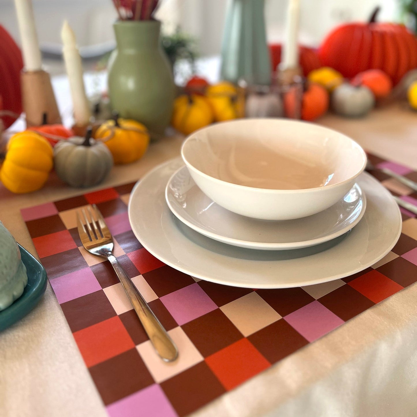 Dining table setting with plates, bowl, and cutlery on a colorful checkered paper placemat with autumn decorations.
