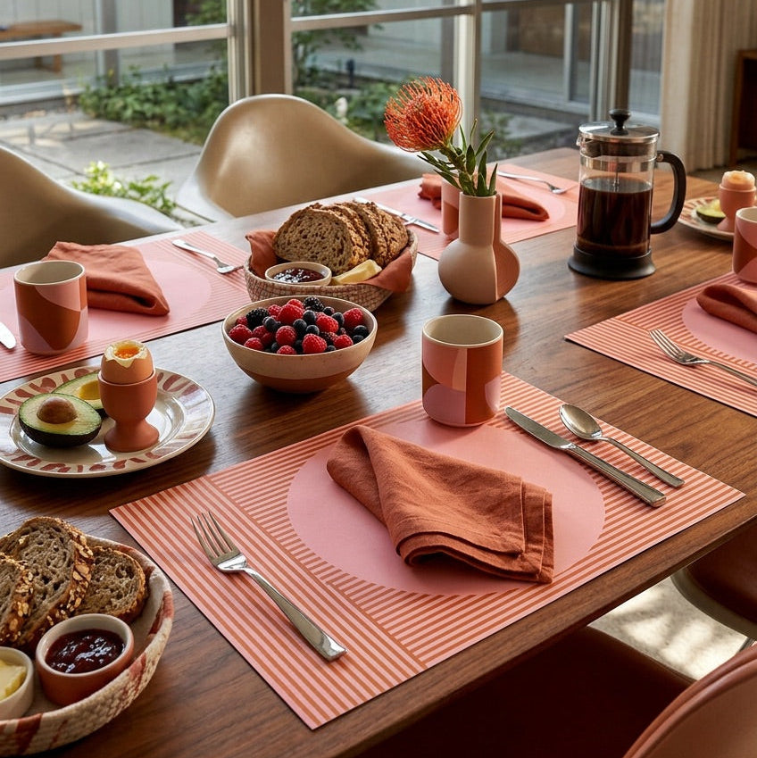 Dining table set with modern striped paper placemats, breakfast items including bread, fruit, and coffee, with a view of a garden through large windows.