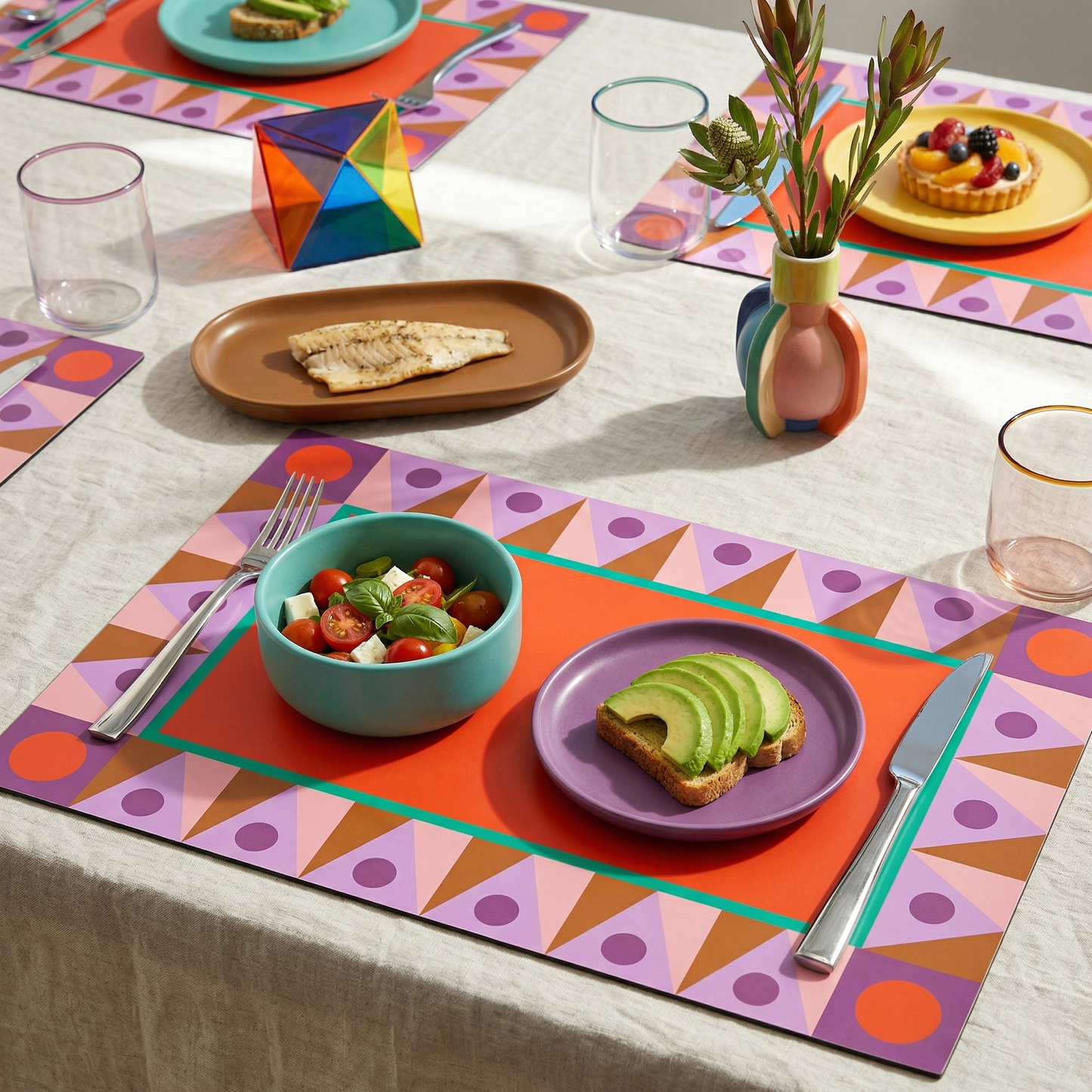 Colorful table setting with geometric paper placemats, plates, and utensils on a white tablecloth.