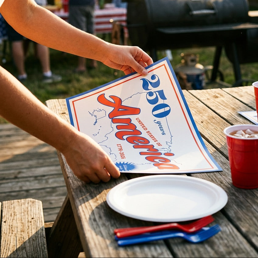 Person holding a 250th America paper placemat at a barbecue event with American flags and people in the background.