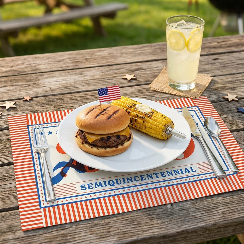 Outdoor setting with a burger, corn on the cob, and lemonade on a 'SemiQuincenennial' paper placemat.