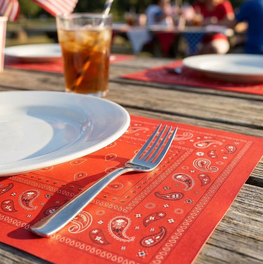 Outdoor setting with plates, glasses, and a fork on a red patterned paper placemat.