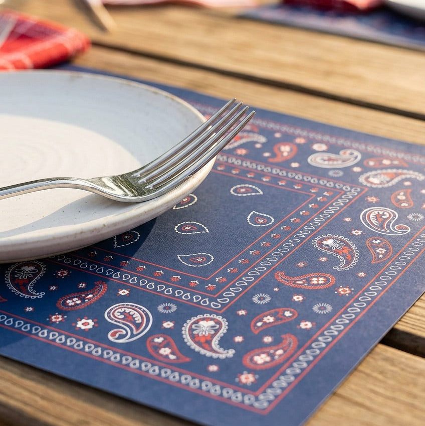 Table setting with blue bandana paper placemats, white plates, and silverware on a wooden table.