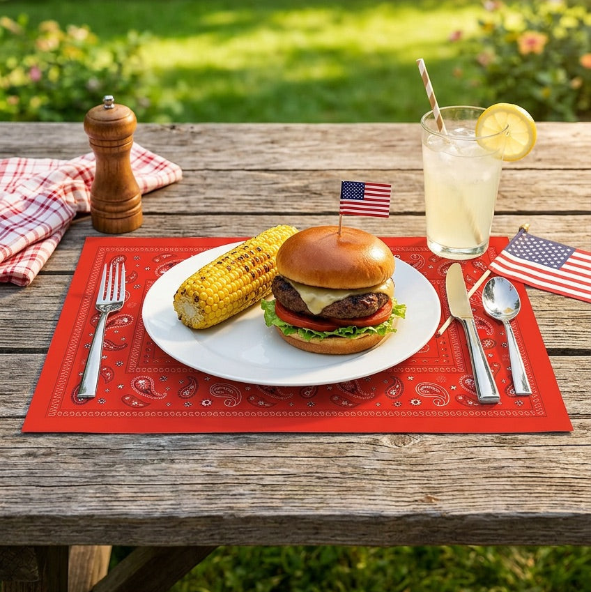 Burger with corn on the cob and a drink on a red paisley paper placemat outdoors.
