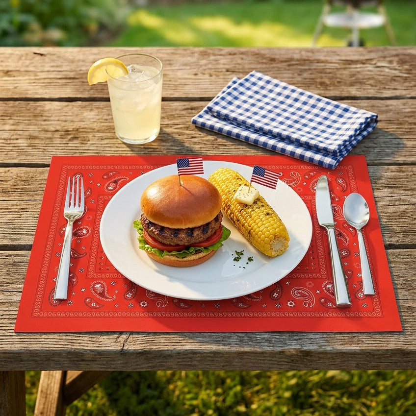 Burger and corn on a plate with a glass of lemonade on a red paper placemat outdoors.