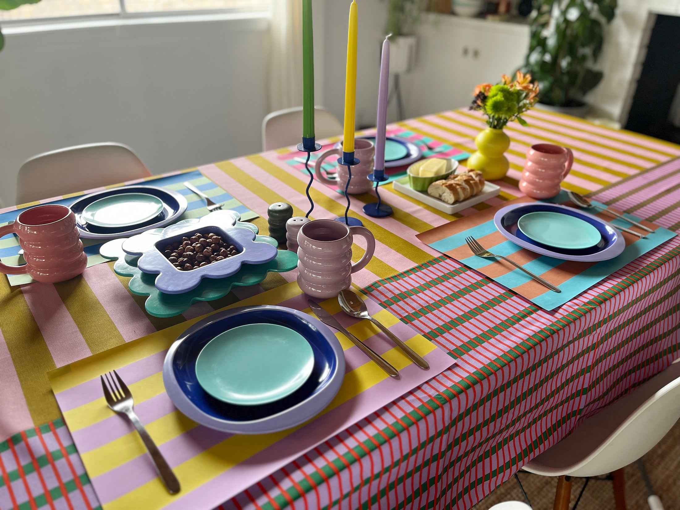 Colorful table setting with plates, mugs, and utensils on a striped tablecloth with striped colorful paper placemats.