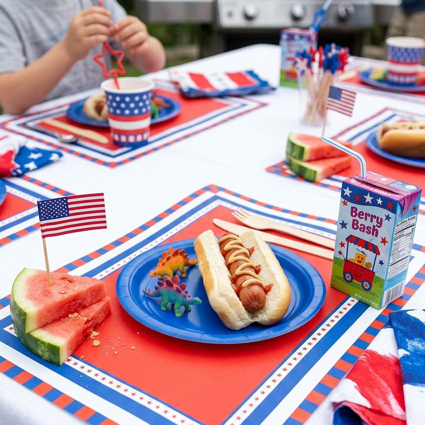 Children's table setting with hot dogs, watermelon, and patriotic placemats on a white tablecloth.