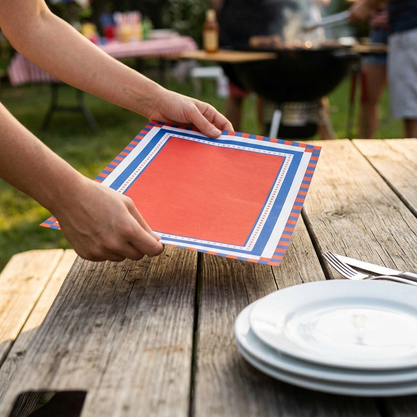 Person setting a table with red and blue placemats outdoors, with a grill and people in the background.
