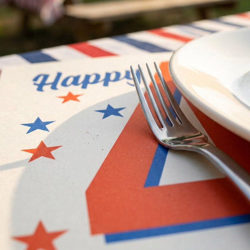 Close-up of a plate with a patriotic paper placemat, fork, and blurred outdoor setting with tables and flags.