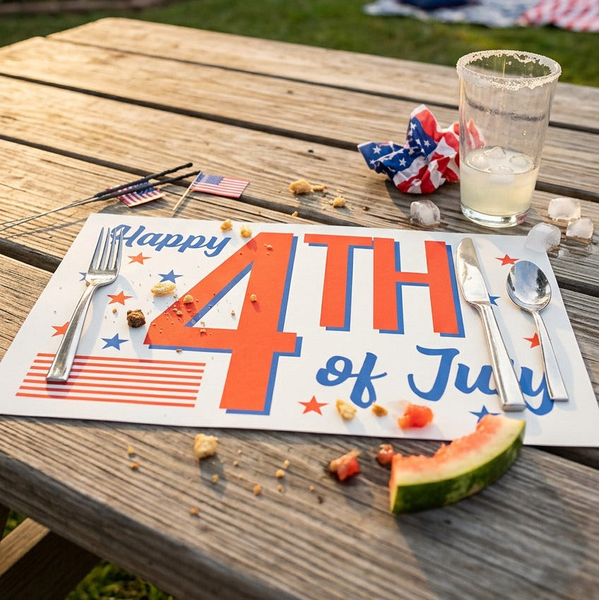 Outdoor table setting with a 'Happy 4th of July' paper placemat, watermelon, and glass of lemonade.