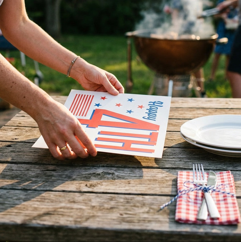 Person setting up a table with a 'Happy 4th of July' paper placemat near a grill and bunting.