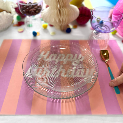 Glass plate with 'Happy Birthday' text on a striped placemat, surrounded by colorful decorations.
