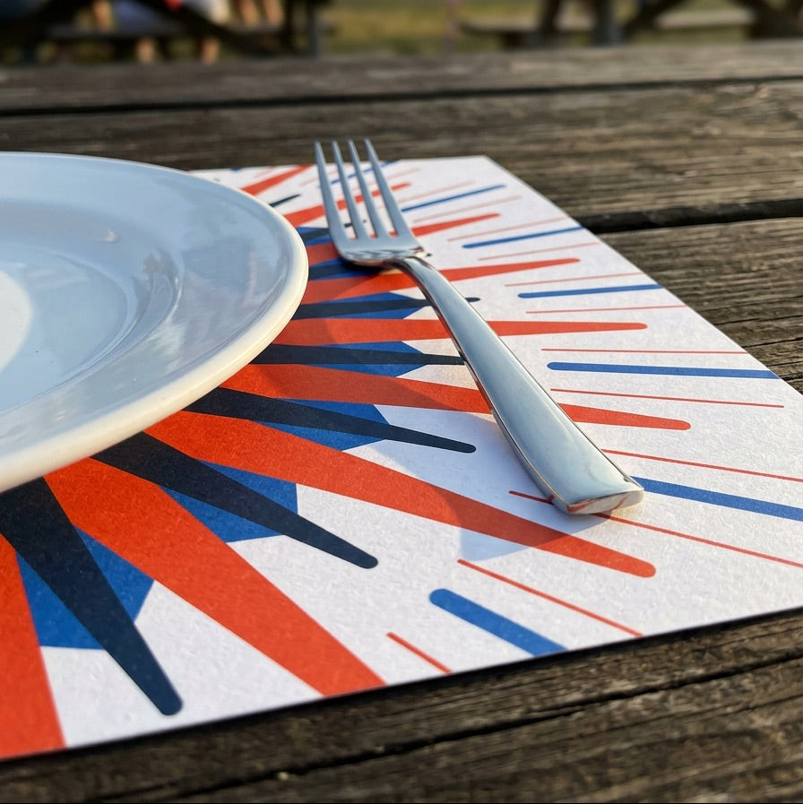Table setting with a plate, fork, and red, white, and blue paper placemat on a wooden table outdoors.