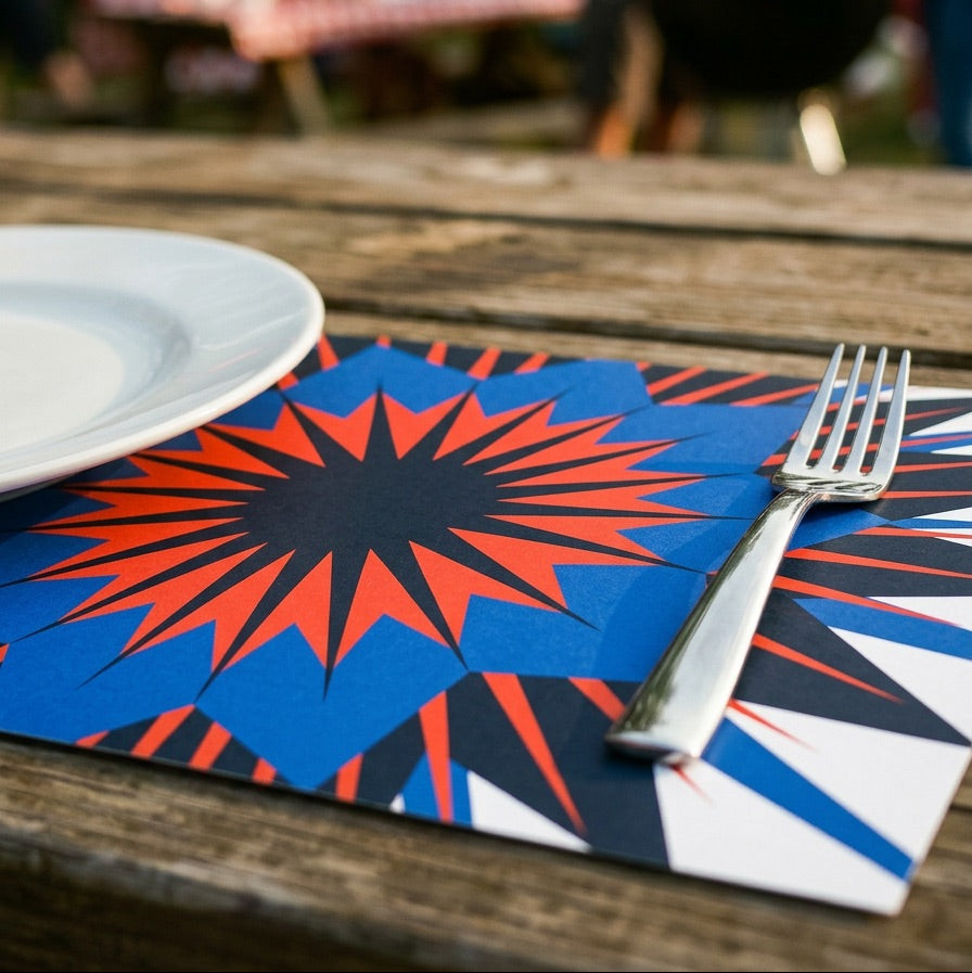 Paper placemat with red and blue star design on a wooden table outdoors, with people and flags in the background.