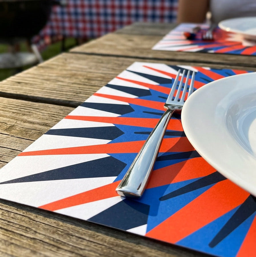 Colorful placemat with a fork and knife on a wooden table outdoors, with people and flags in the background.
