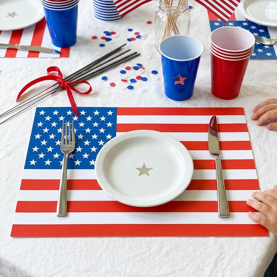 Table setting with American flag paper placemats, cups, and cutlery on a white tablecloth.