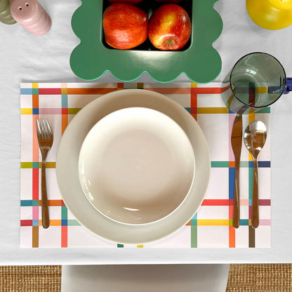 Dinner setting with a colorful paper placemat, white bowl, cutlery, and fruit on a white table.