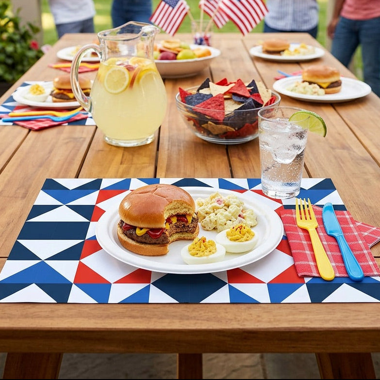 Outdoor gathering with a table set on patriotic paper placemats, featuring burgers, chips, and drinks, with American flags in the background.