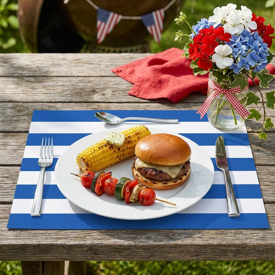 Outdoor setting with a grill, plate of food, and flowers on a blue and white striped paper placemat.