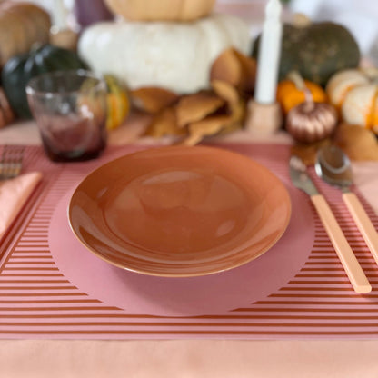 Table setting with a terracotta plate on a pink striped paper placemat, surrounded by pumpkins and autumn decorations.