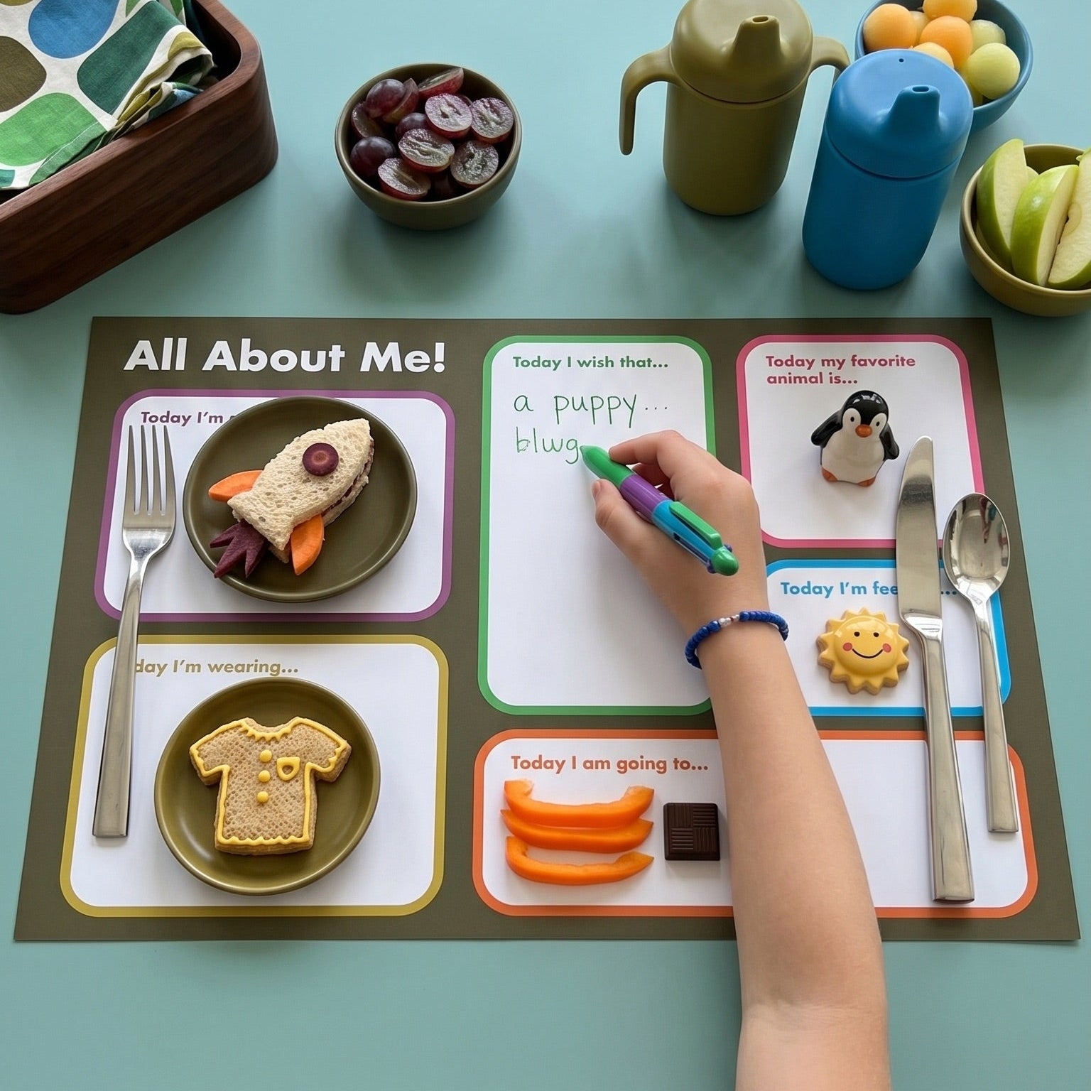Child's hand filling out a 'All About Me' worksheet on a table with food and utensils.