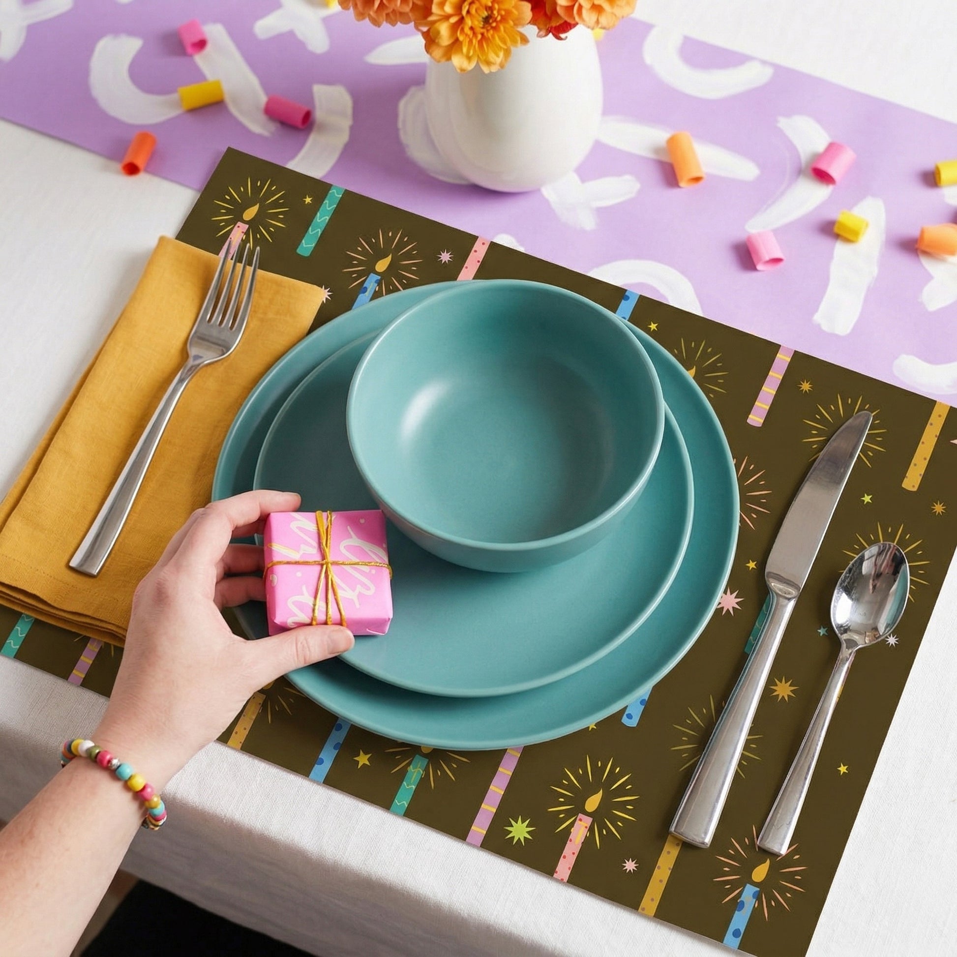 Teal dinnerware set on a colorful placemat with a hand holding a gift box.