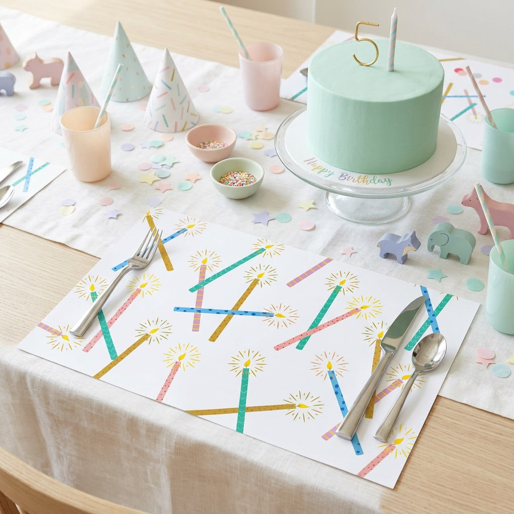 Table setting with a birthday cake, colorful paper placemats with candles, and utensils on a wooden table.