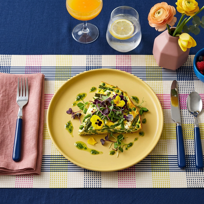 Dinner setting with a plate of food, cutlery, and a colorful paper placemat on a blue tablecloth.