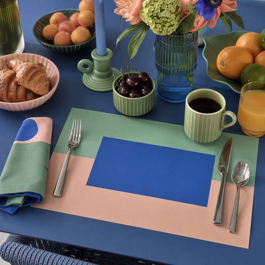 Colorful table setting with a blue and pink paper placemat, cutlery, and fruit on a blue tablecloth.