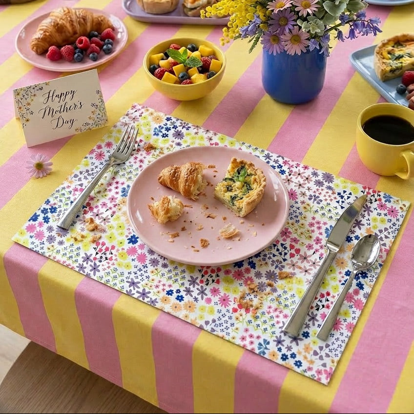 Dining table set for breakfast with pastries, fruit, coffee, and flowers on a colorful paper placemat.