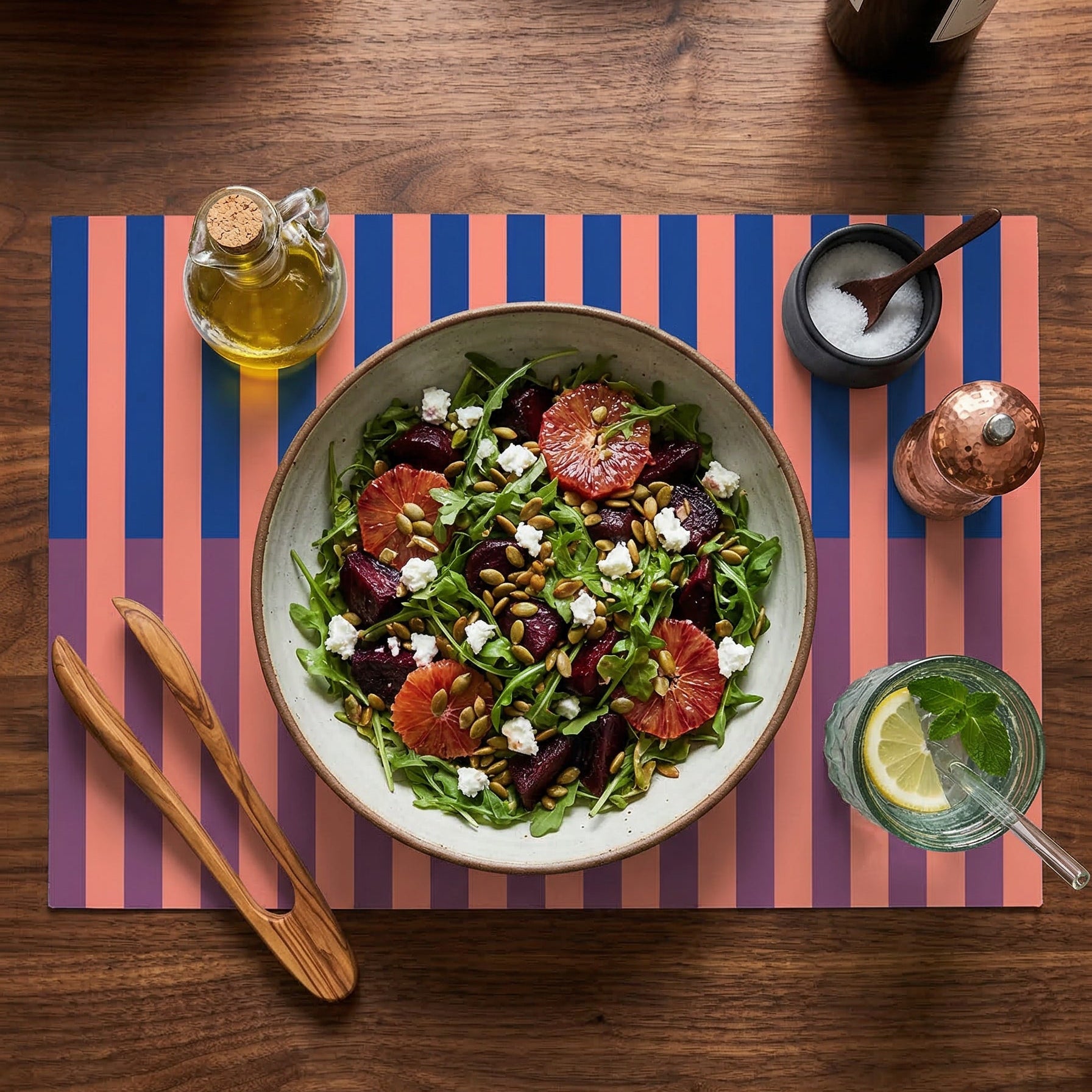 Salad on a striped paper placemat with olive oil, salt, and a bottle of wine on a wooden table.