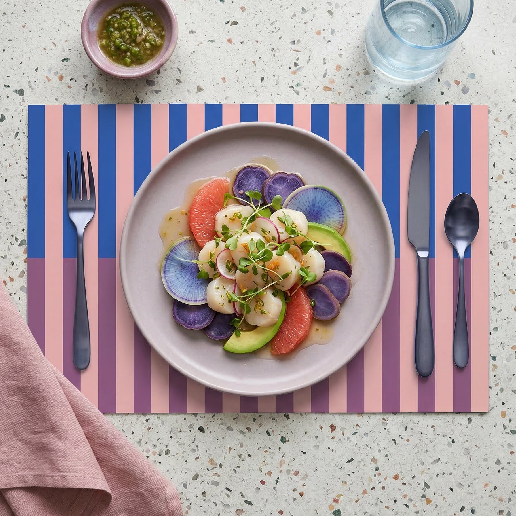 Striped paper placemat with a plate of salad, cutlery, and a glass of water on a speckled countertop.