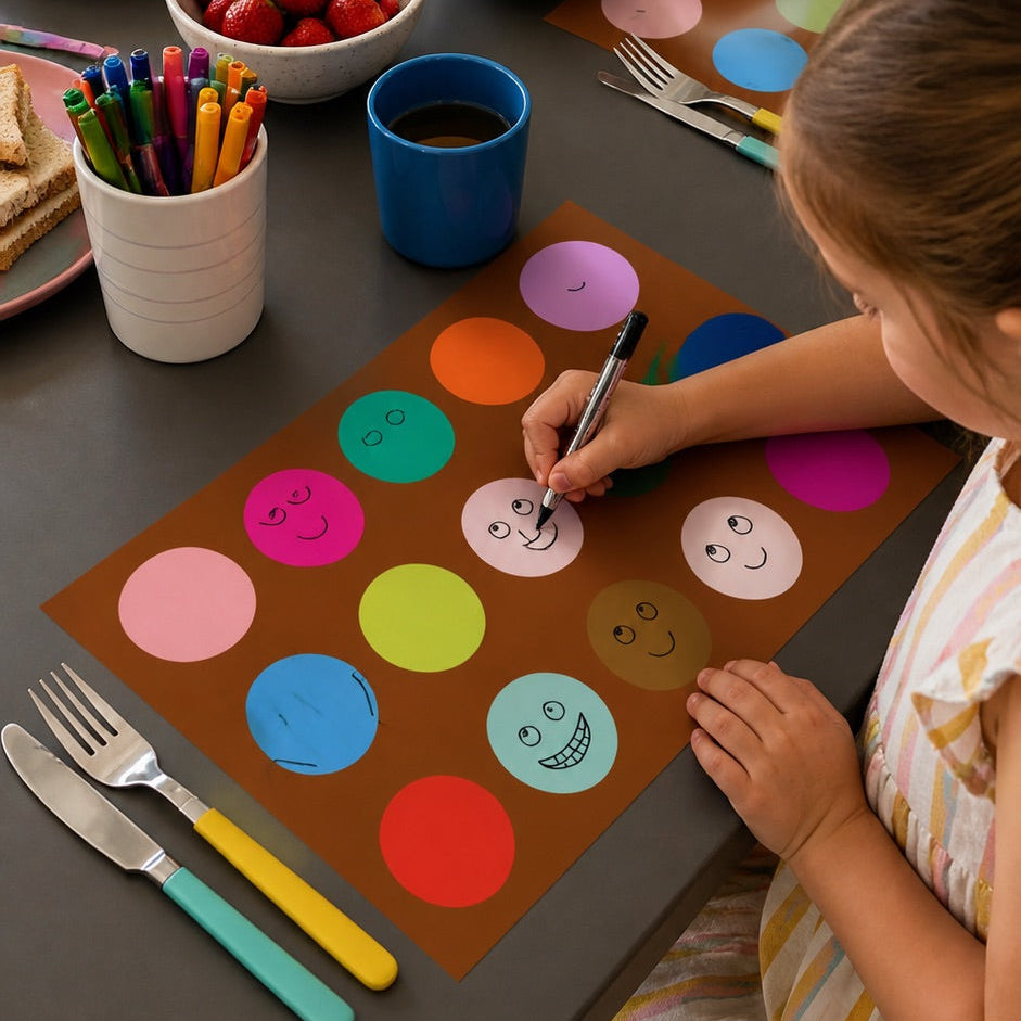 Child coloring on a placemat at a dining table with snacks and utensils.