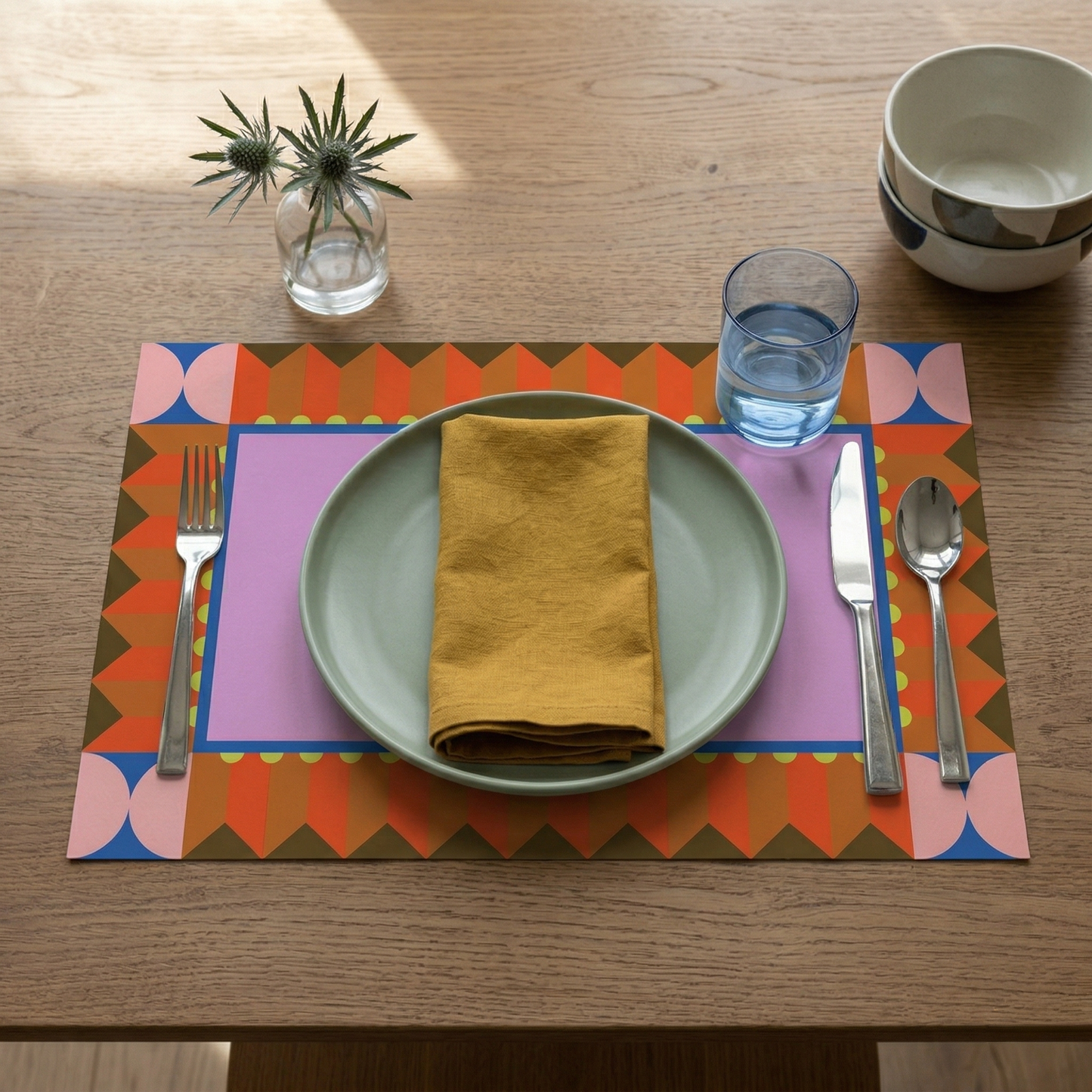 Dinner setting with a colorful placemat, plate, fork, knife, spoon, and glass on a wooden table.