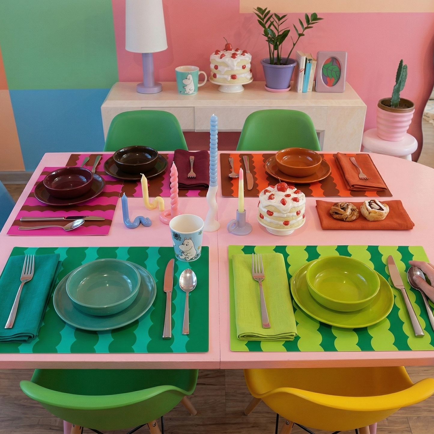 Children's play table with colorful placemats, plates, and chairs in a room with a multicolored wall.