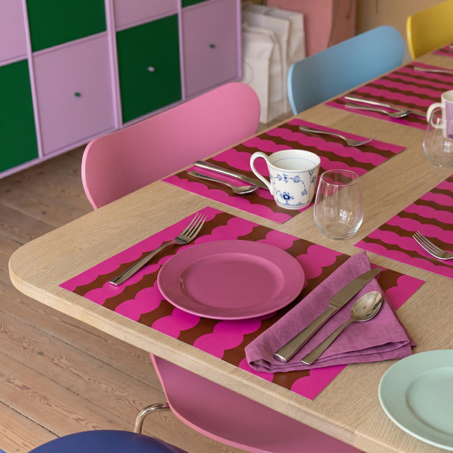 Children's table setting with pink plates, cutlery, and paper placemats in a colorful room.