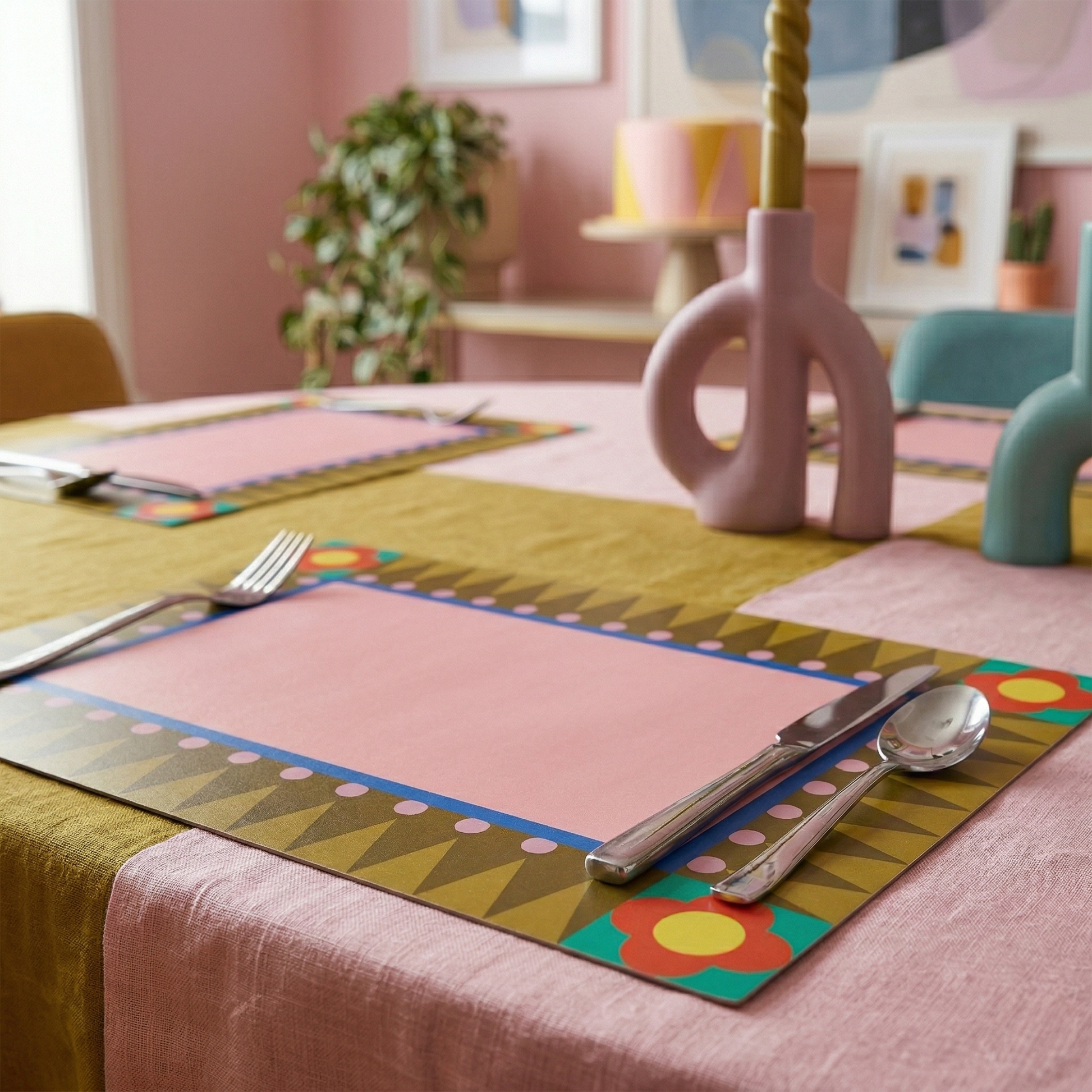 Colorful geometric paper placemats with cutlery on a table in a room with pink walls and plants.