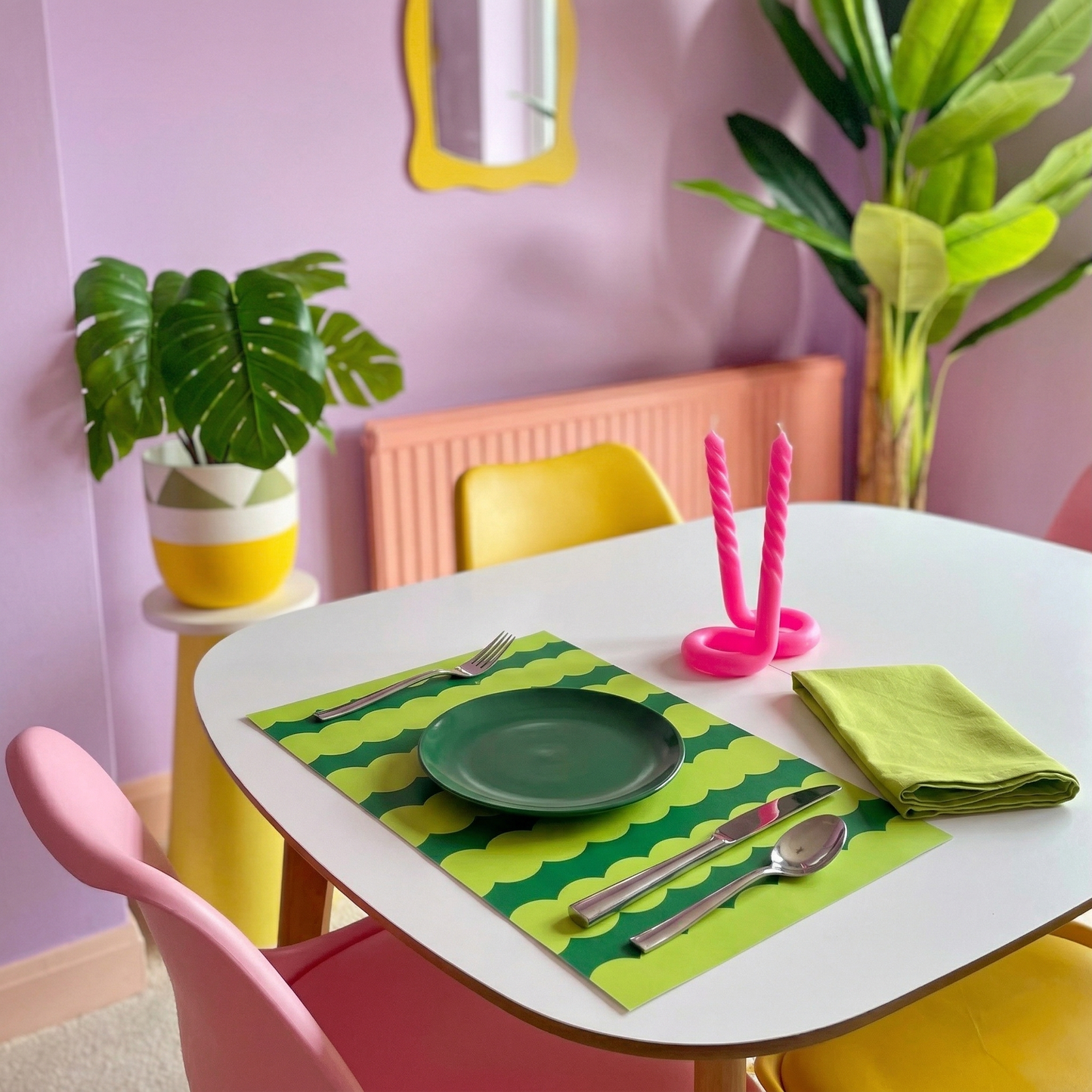 Dining table setting with green plate, napkin, and green wavy striped paper placemat in a room with pink walls and plants.