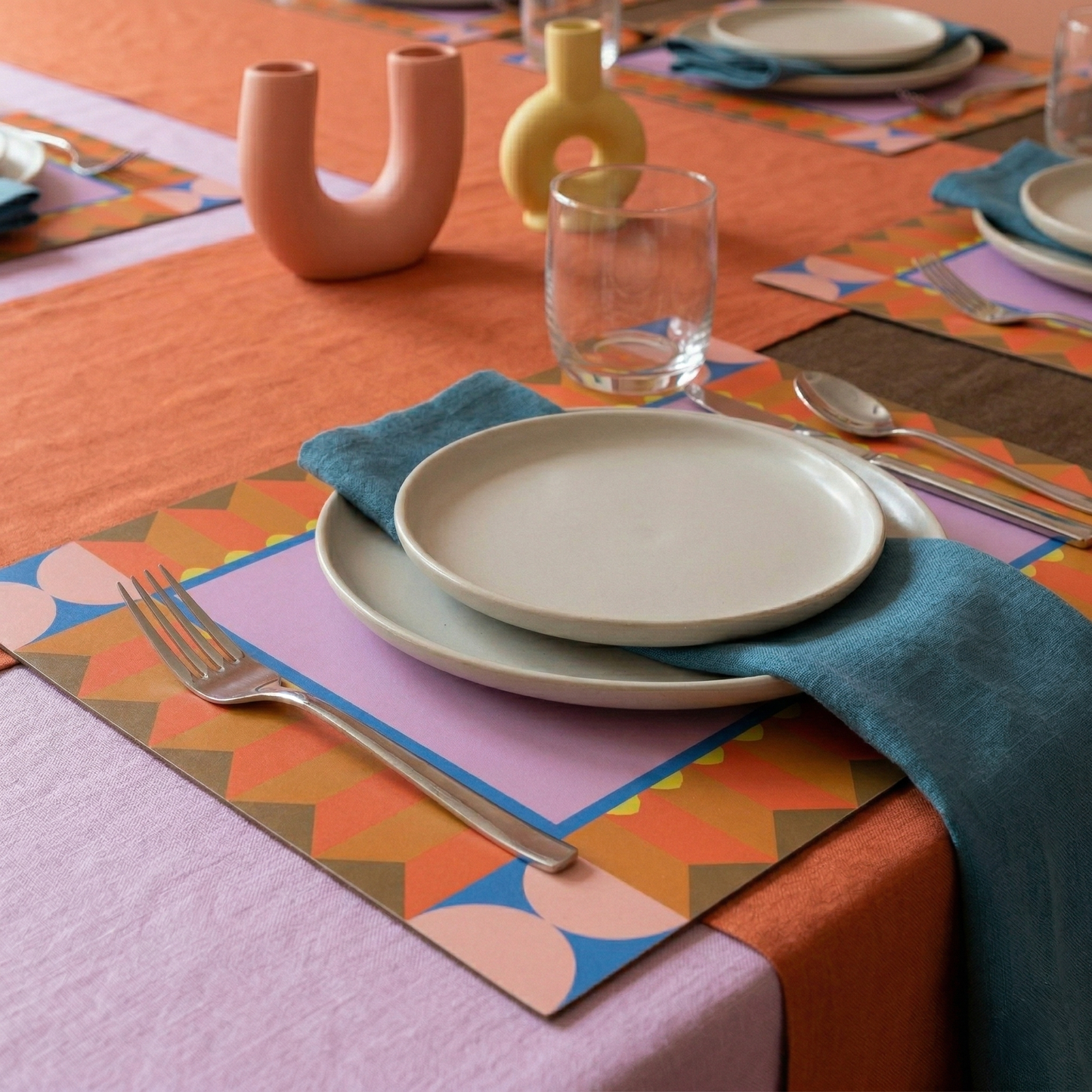 Colorful table setting with geometric placemats, plates, and cutlery on a patterned tablecloth.
