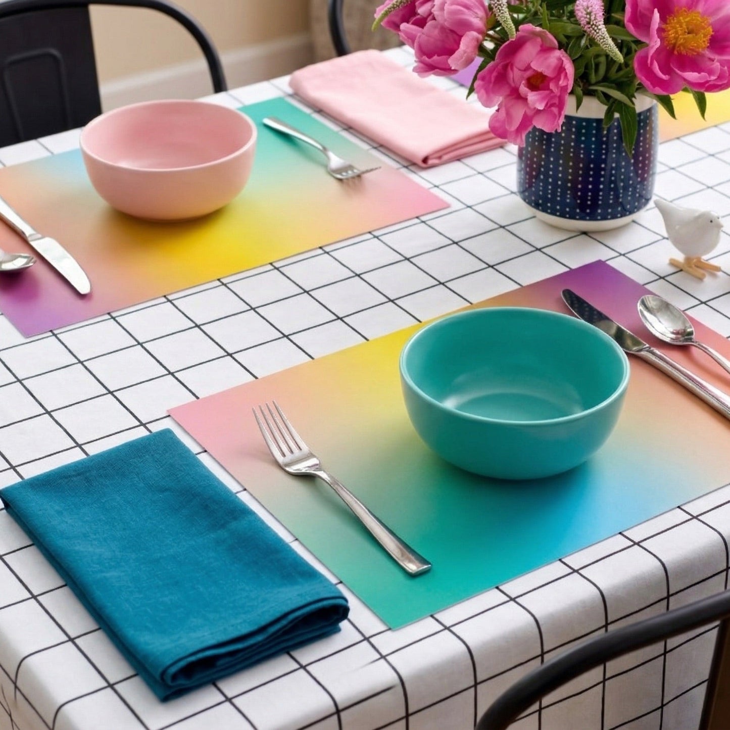 Dining table with colorful paper placemats, bowls, and cutlery on a checkered tablecloth.