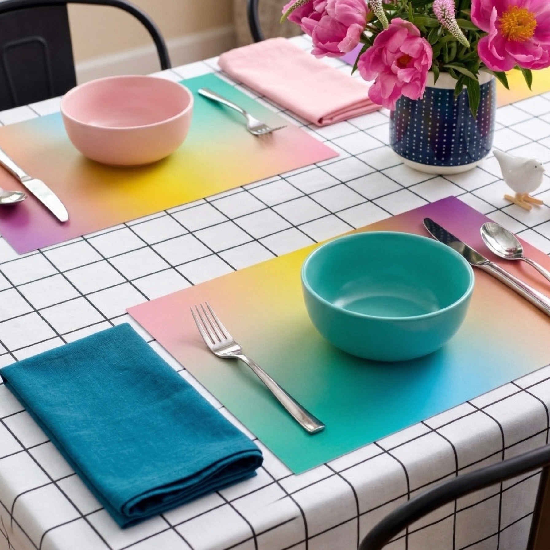 Dining table with colorful paper placemats, bowls, and cutlery on a checkered tablecloth.