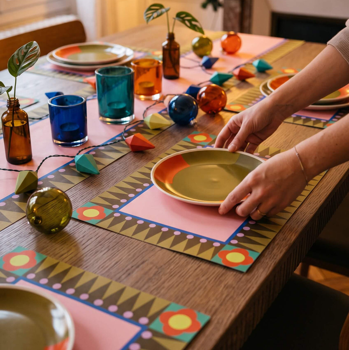 A Christmas lifestyle shot of a dining table decorated for a holiday gathering, featuring colorful geometric placemats, matching plates, amber glass bottles with greenery, and string lights. A person's hands are visible placing a plate on one of the placemats.