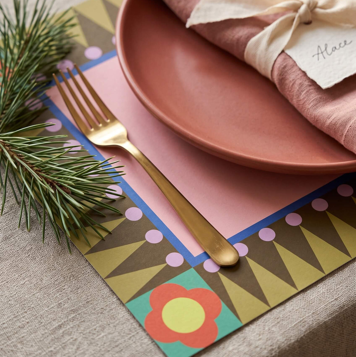 A styled holiday table setting featuring a geometric paper placemat with retro patterns, a dusty rose ceramic plate, a gold fork, and a pink linen napkin tied with a cream ribbon and a paper tag.
