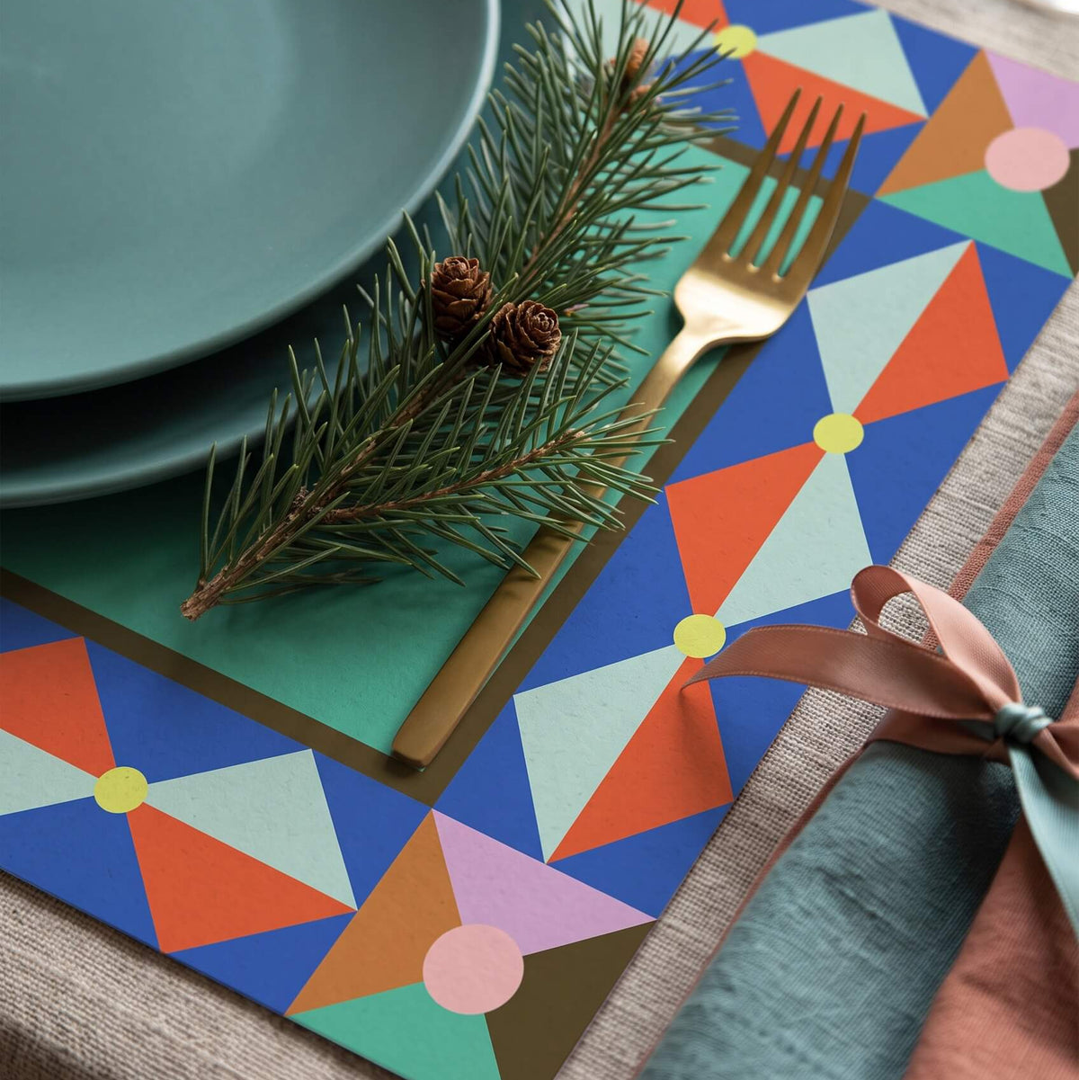 A styled Christmas table setting featuring a colorful geometric placemat with blue, orange, green, and pink triangles. The placemat is styled with a teal ceramic plate, a gold fork, a pine branch with pinecones, and a folded teal napkin with a dusty rose ribbon on a light wood table.
