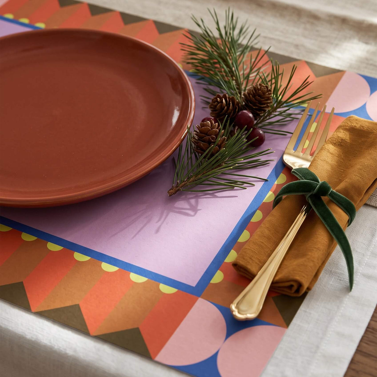 A styled Christmas table setting featuring a multicolor geometric placemat with orange, pink, blue, and purple shapes, a terracotta plate, a gold fork on a mustard napkin tied with a green ribbon, and pine sprigs on a beige table runner.