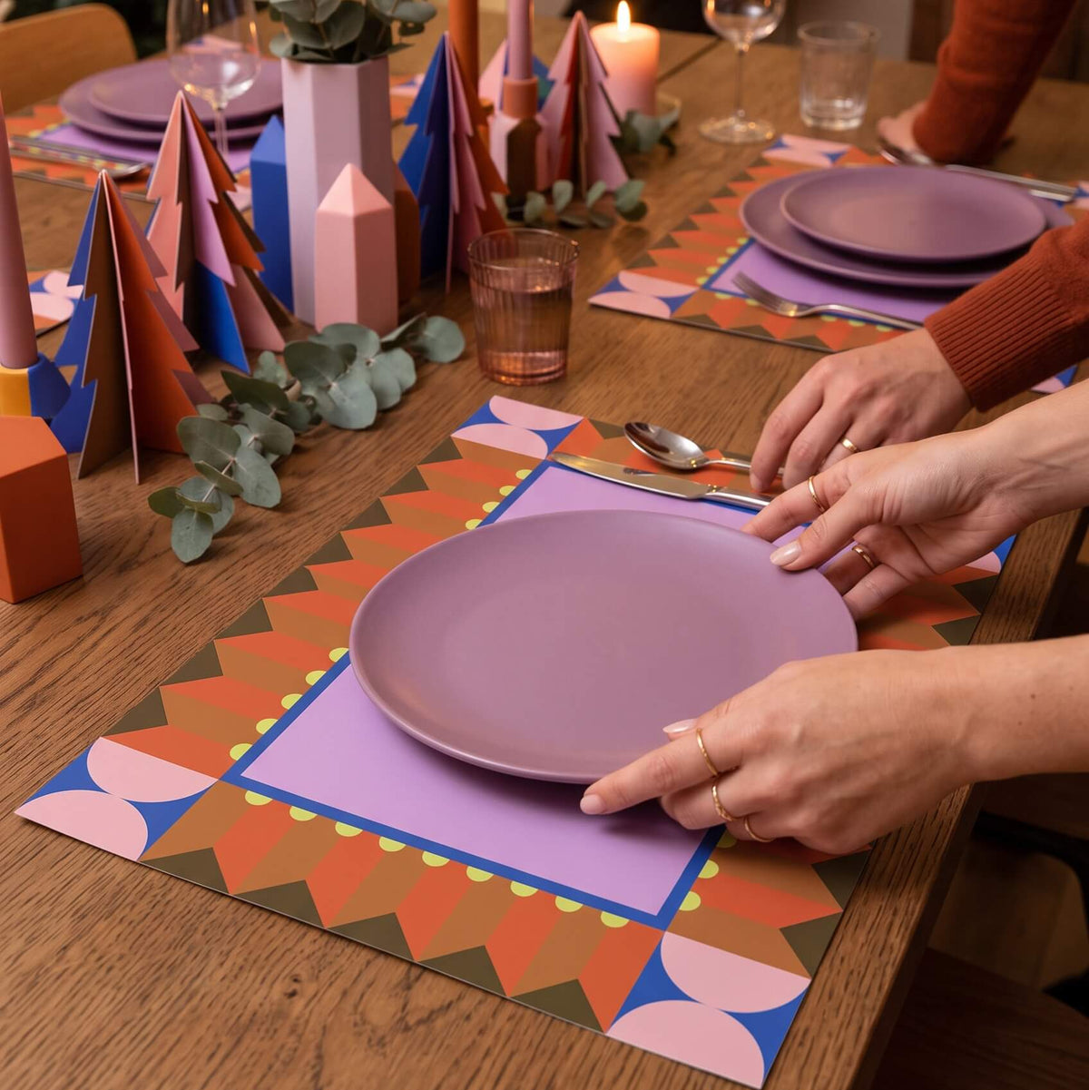 A styled dining table set with colorful geometric placemats and matching purple plates, decorated with candles, eucalyptus branches, and paper trees in a warm, festive indoor setting. A person's hands are shown placing a purple plate on a placemat in the foreground.